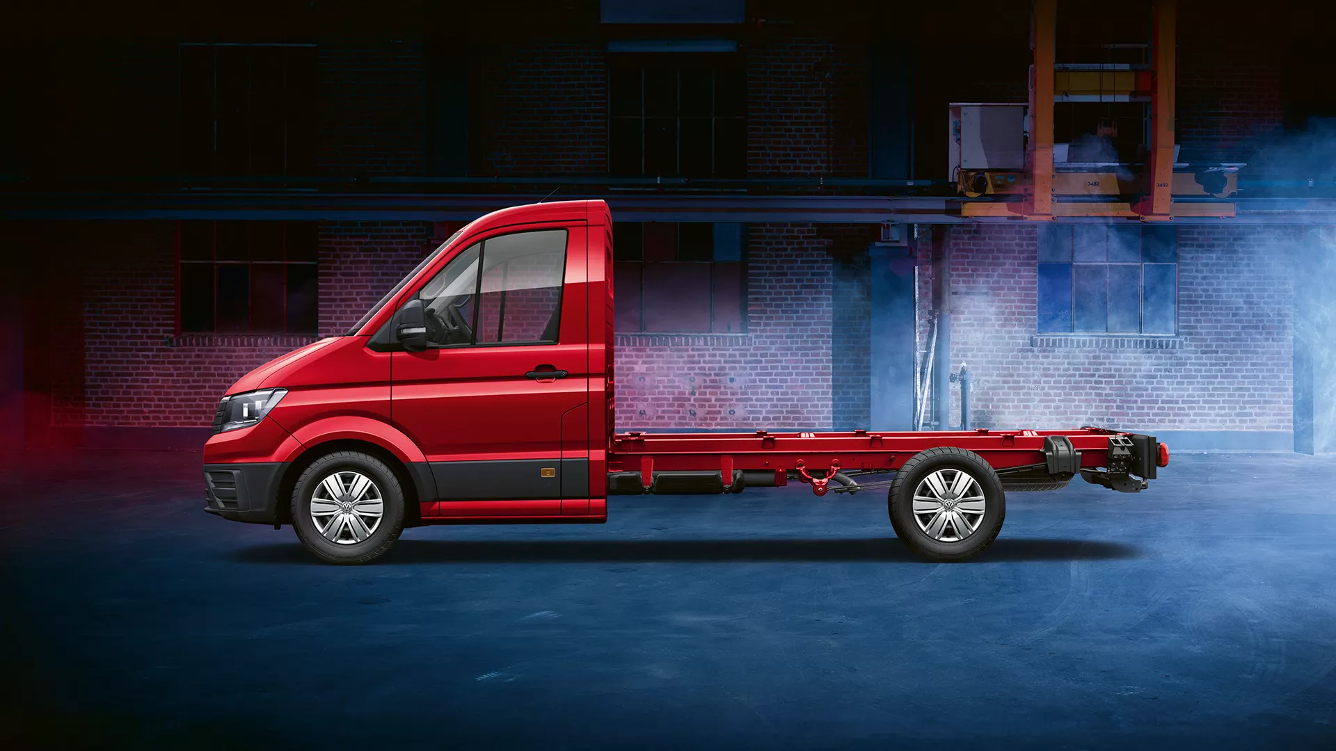 Camión eléctrico Volkswagen Crafter Chasis de color rojo brillante, estacionado en una calle urbana con edificios al fondo envueltos en humo azul, mostrando la potencia y tecnología del vehículo.