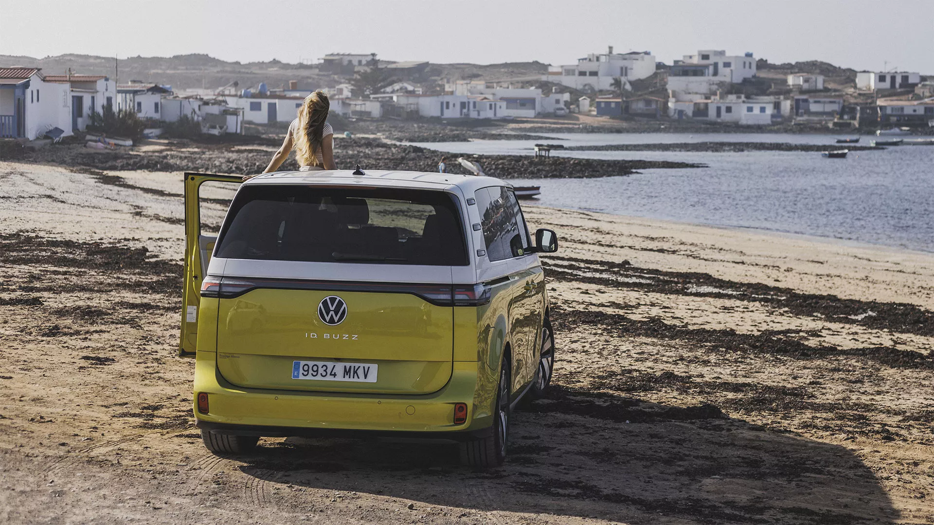 Un automóvil Volkswagen ID Buzz de color verde claro estacionado en la playa, con una persona sentada en el techo mientras se observa una vista panorámica de un pueblo costero en segundo plano.