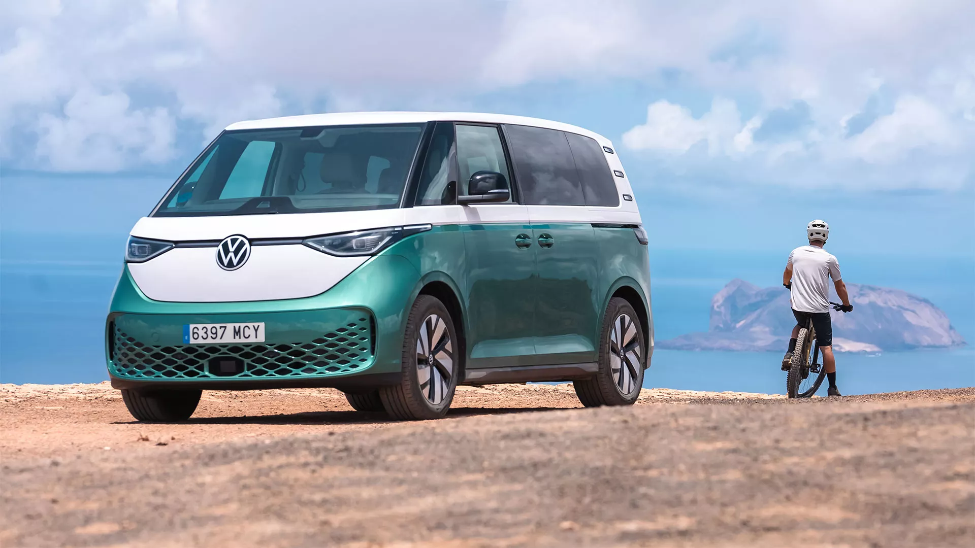 Vehículo eléctrico Volkswagen ID.Buzz de color blanco y verde estacionado en un escenario montañoso y costero, con una persona con bicicleta junto a él