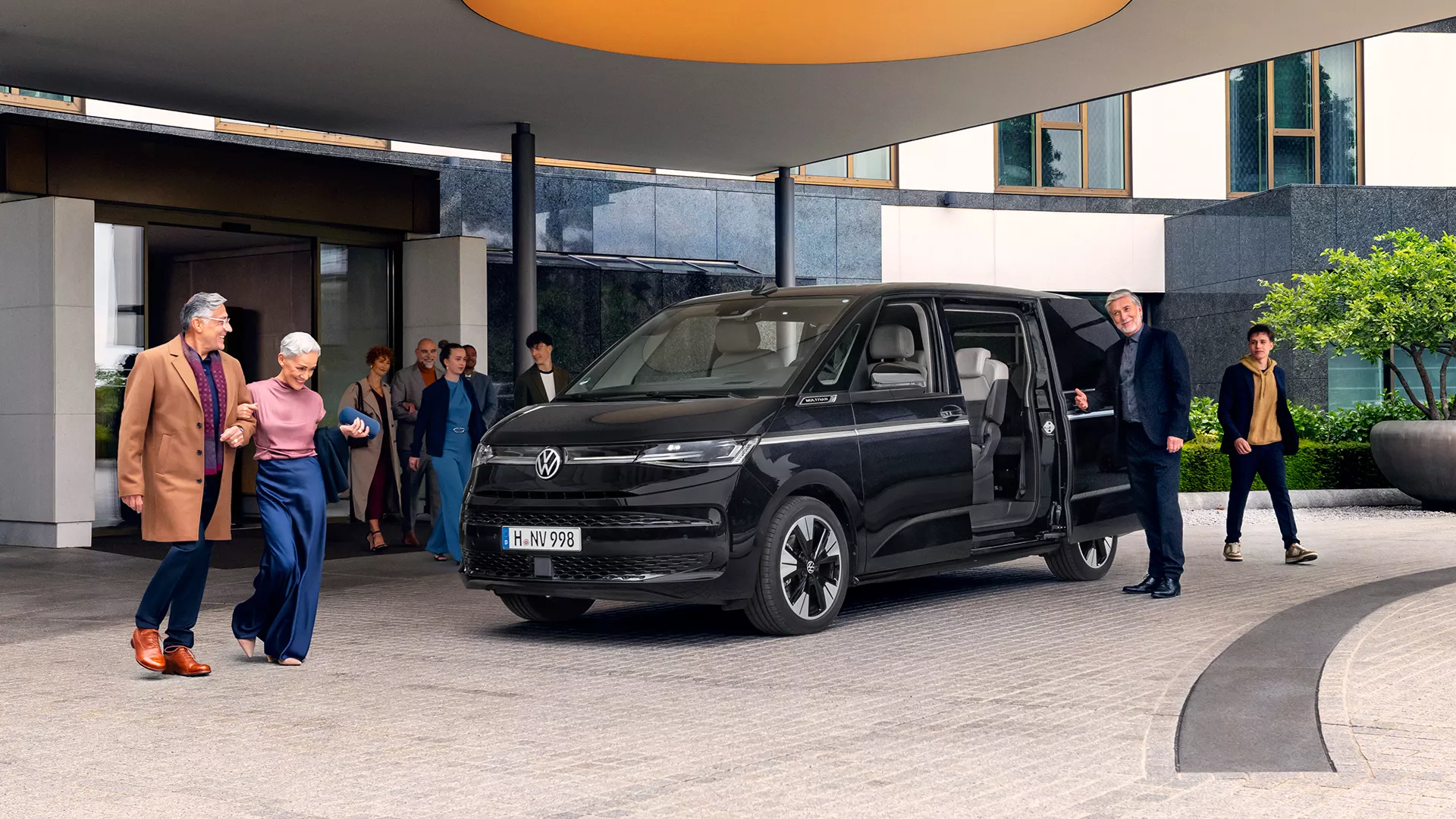 Un grupo de personas elegantemente vestidas observa un auto Volkswagen Multivan de color negro aparcado frente a un edificio moderno con grandes ventanales.