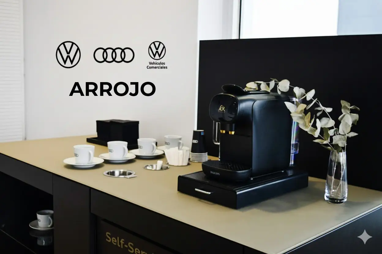 Mostrador de recepción con logo de Volkswagen y Audi, equipado con una cafetera, tazas y platos, y un arreglo floral. La imagen representa una zona de descanso o de bienvenida en una concesionaria de Vehiculos Comerciales.