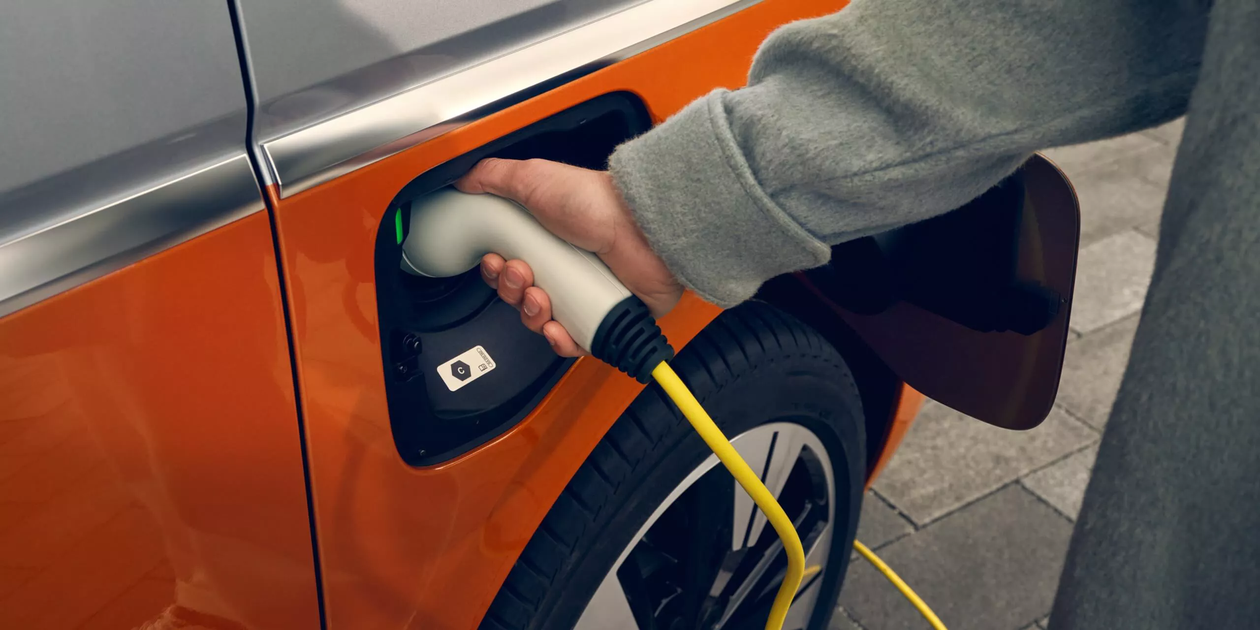 La imagen muestra un cable de carga eléctrica conectado a la toma de un vehículo Volkswagen Multivan eléctrico, evidenciando la transición hacia la movilidad sostenible.