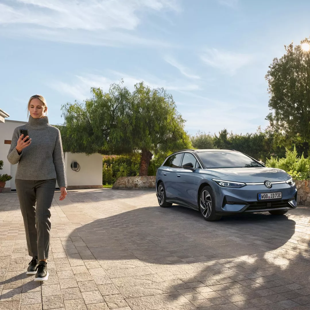 Una persona en un jersey gris de cuello alto camina por una plaza de ladrillos junto a un automóvil Volkswagen eléctrico estacionado, con un paisaje verde y árboles en segundo plano.