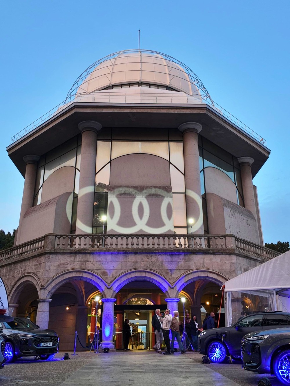 Edificio circular con iluminación morada y coches Arro en la entrada de un evento nocturno